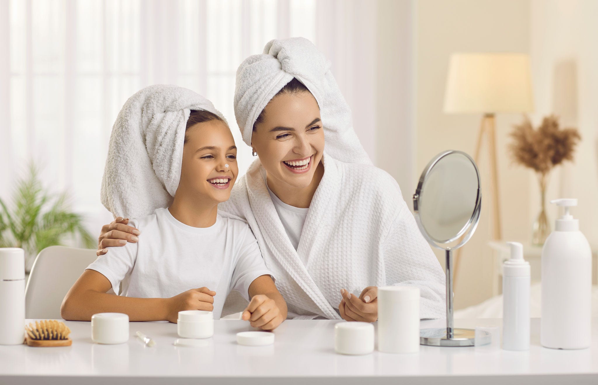 Happy mother and child enjoying morning skin care routine together. Cheerful beautiful mommy and little daughter looking in small mirror on beauty table in bedroom, applying face cream and laughing