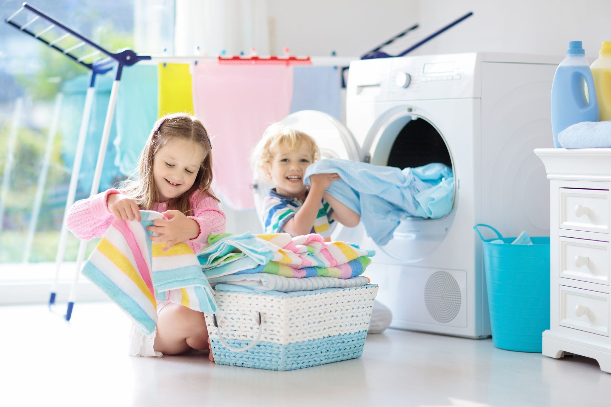Children in laundry room with washing machine or tumble dryer. Kids help with family chores. Modern household devices and washing detergent in white sunny home. Clean washed clothes on drying rack.